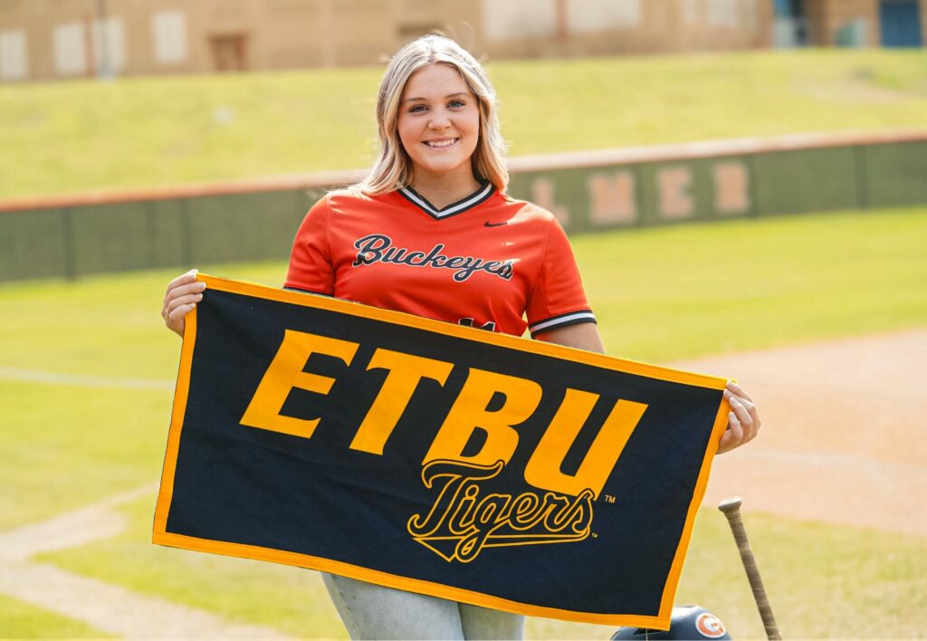 A teenage blond girl smiling and posing on a baseball field, wearing an orange Buckeyes team shirt and holding a blue and yellow banner with ETBU Tigers written on it | Daniel Stark Law