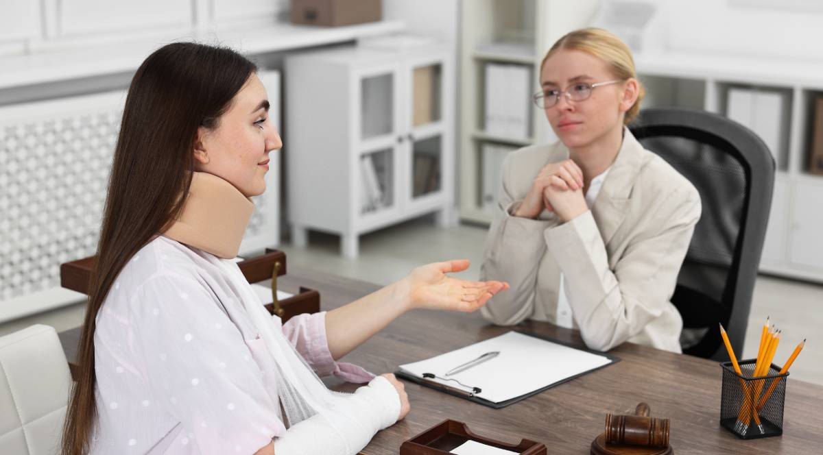 Dark-haired woman in a neck brace and arm cast meeting with an attorney in a bright, modern office | Daniel Stark Injury Lawyers