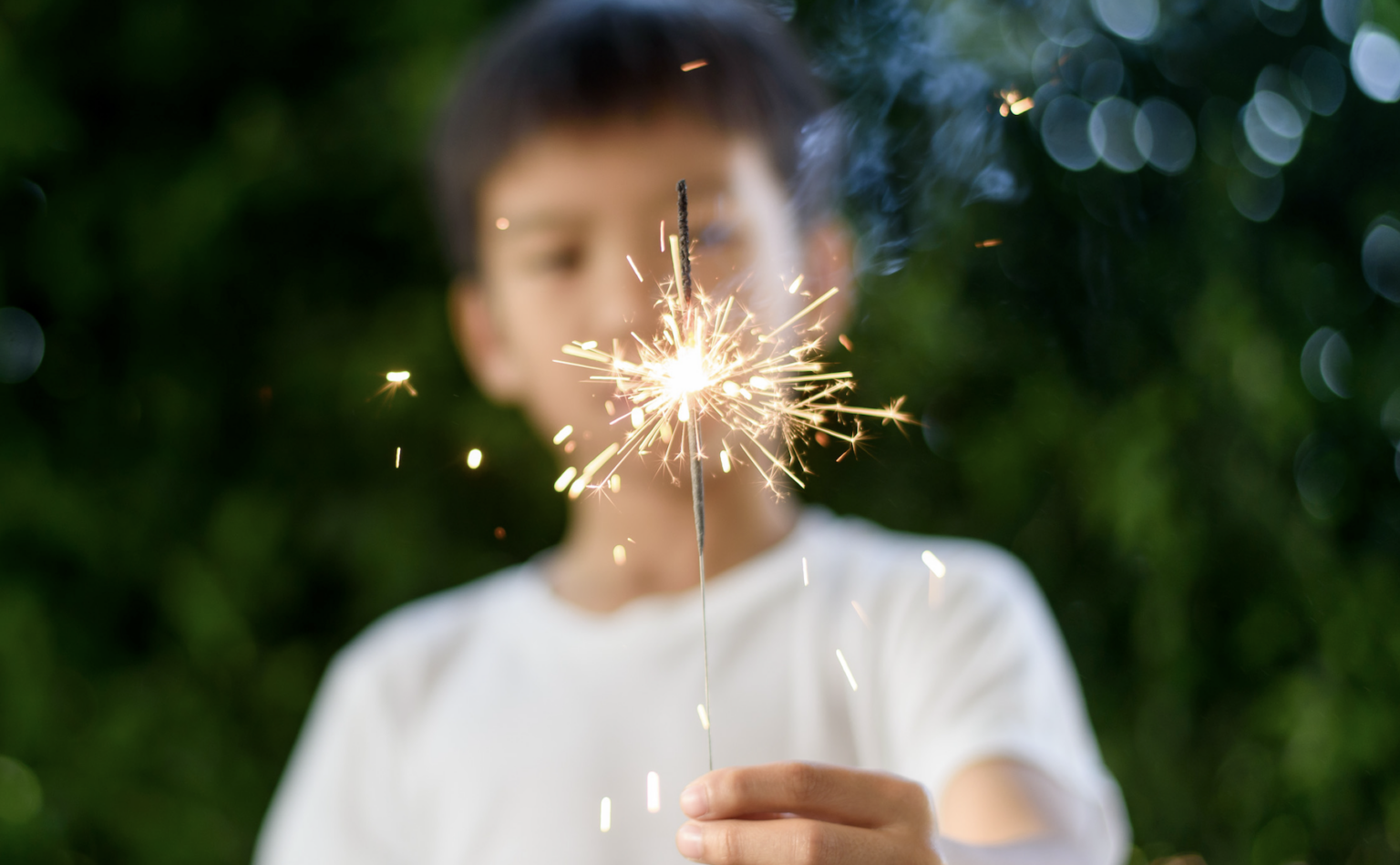 Blurred image of a young boy holding a burning sparkler, with the bright sparkler flame in focus during a fireworks celebration | Daniel Stark Injury Lawyers