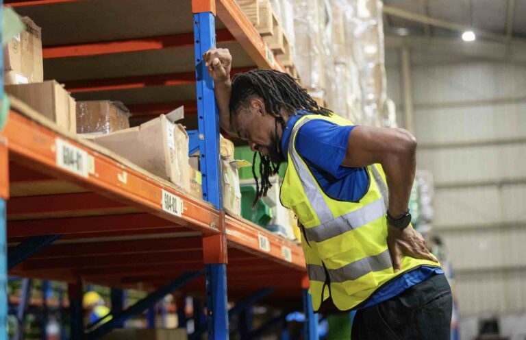 Male warehouse worker holding his lower back after a workplace injury while lifting boxes | Daniel Stark Injury Lawyers