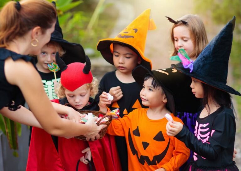 Group of children dressed in Halloween costumes gathered around a woman holding a bucket of candy during daytime trick-or-treating | Daniel Stark Injury Lawyers